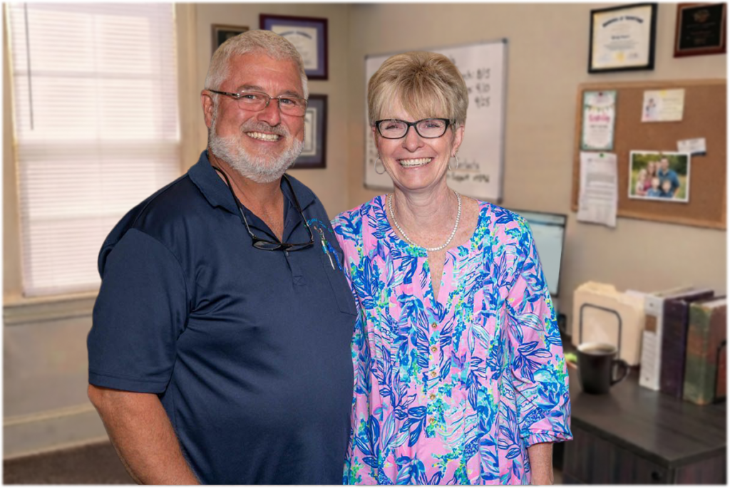 Two smiling adults stand together in an office, with certificates and documents visible on the wall and desk behind them.