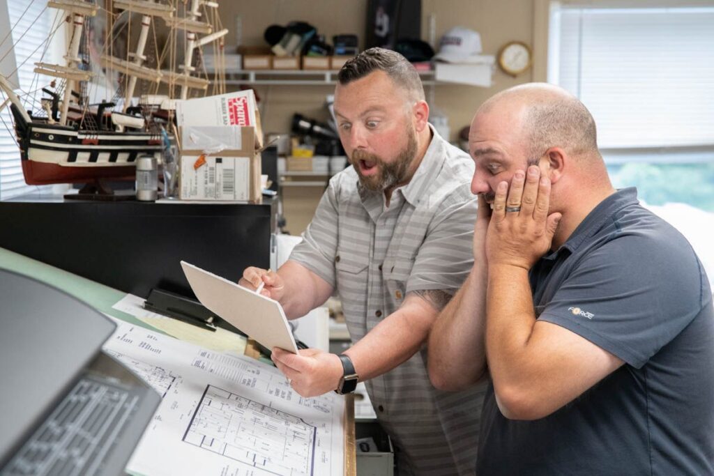Two men in an office look surprised while reading a document; one points at the paper, and the other holds his face. Blueprints and a model ship are visible on the desk.