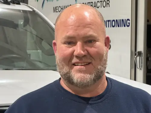 A man with a shaved head and trimmed beard is standing and smiling in front of a white van with business signage visible in the background.